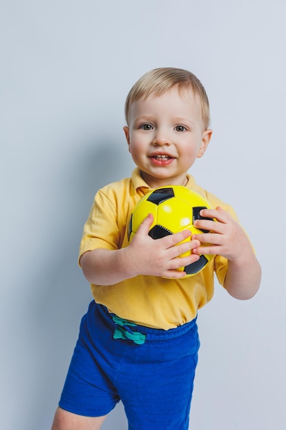 Um garotinho com uma bola de futebol nas mãos em um fundo branco uma criança é um jogador de futebol iniciante um esporte para crianças Atleta pequeno Kit de futebol amarelo e azul para crianças