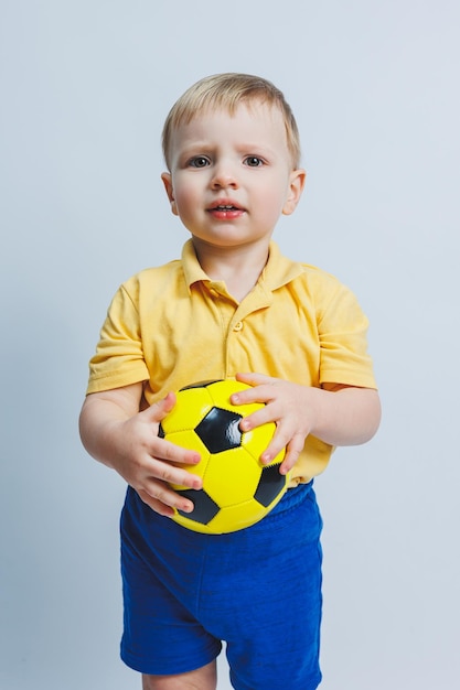 Foto um garotinho com uma bola de futebol nas mãos em um fundo branco uma criança é um jogador de futebol iniciante um esporte para crianças atleta pequeno kit de futebol amarelo e azul para crianças