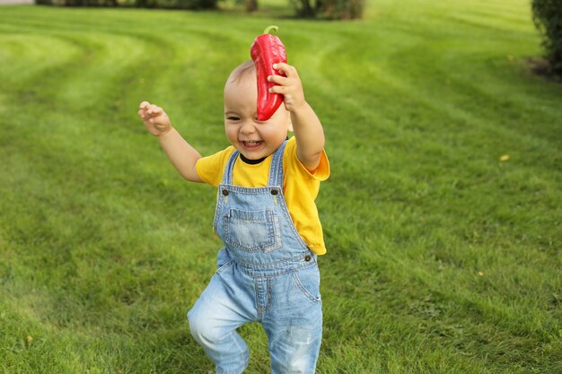 um garotinho com uma camiseta amarela está parado no gramado do parque segurando um pimentão vermelho