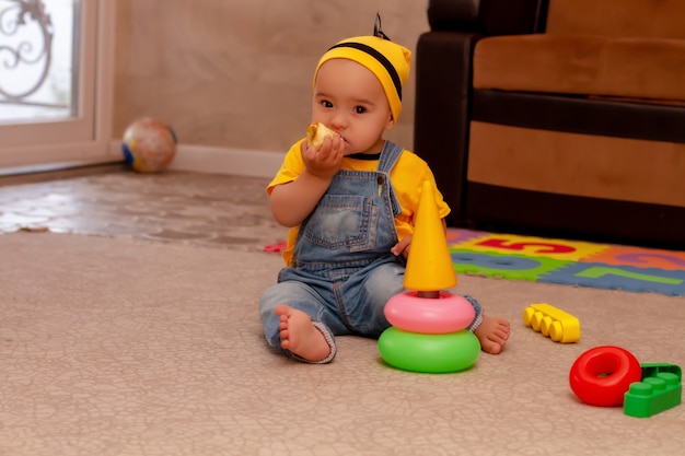 Foto um garotinho com uma camiseta amarela, um chapéu e um macacão jeans está sentado em uma sala em casa