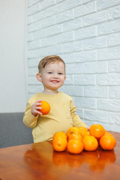 Um garotinho de 2 anos está de pé em uma mesa com tangerinas Bebê quer sentar frutas cítricas pela primeira vez