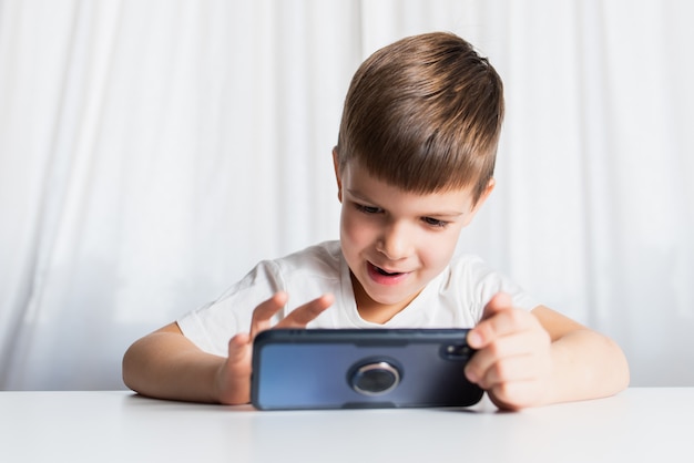 Um garotinho de camiseta branca joga em casa ao telefone. Uma criança feliz olha para seu smartphone.
