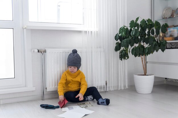 Um garotinho de suéter amarelo e chapéu está contando dinheiro e estudando contas de aquecimento perto de um aquecedor com termostato