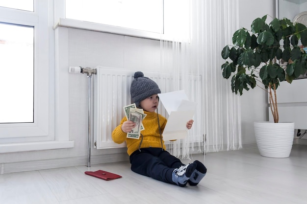 Um garotinho de suéter amarelo e chapéu está contando dinheiro e estudando contas de aquecimento perto de um aquecedor com termostato