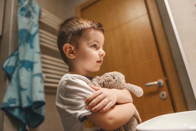 Um garotinho está parado no banheiro com um coelho nas mãos e se olhando no espelho