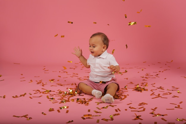 um garotinho feliz em uma camiseta branca e shorts vermelhos está sentado sobre um fundo de confete rosa