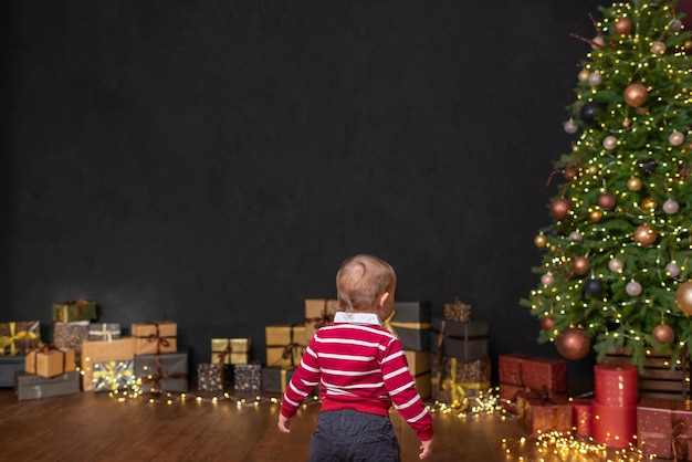 Um garotinho fica em frente a uma árvore de Natal e caixas com presentes ao longo de uma parede preta