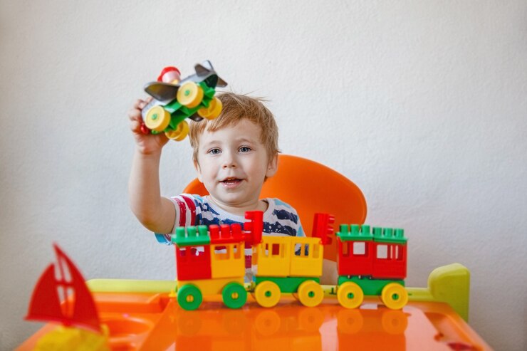 Um garotinho loiro está sentado em casa em sua mesa entre brinquedos de plástico brilhantes e brinca com o avião segurando-o. Foto de alta qualidade