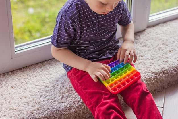 Um garotinho loiro garotinho loiro brincando com um brinquedo popit moderno colorido e brilhante covinha simples. A criança com um colorido brinquedo sensorial de silicone antiestresse