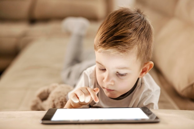 Um garotinho no sofá na sala está lendo ebook no tablet ele está rolando as páginas