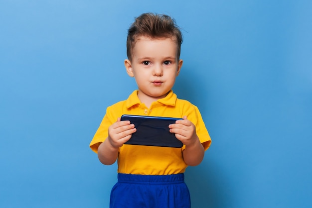 Um garotinho olha para a tela de um telefone celular. foto em uma parede azul.