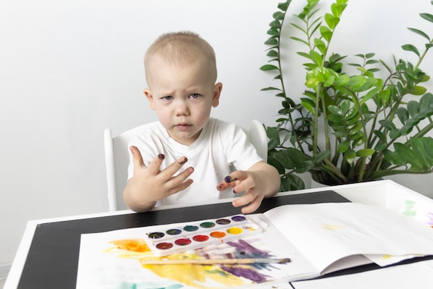 Um garotinho pinta com tintas aquarela em uma mesa ao lado de uma flor verde