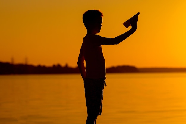 Um garotinho tem um avião de papel na mão no fundo do rio ao pôr do sol A silhueta do menino com origami na mão Linda paisagem noturna