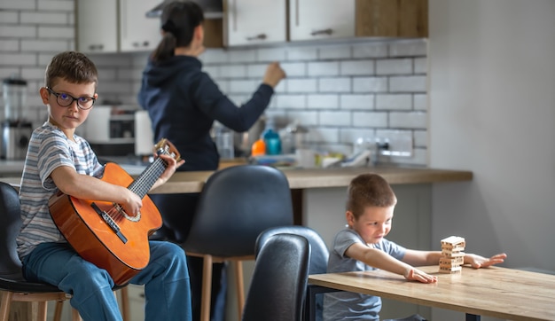 Foto um garotinho toca violão, e seu irmão constrói uma torre com cubos de madeira em casa na mesa.