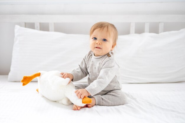 Foto um garotinho, uma criança loira de olhos azuis, de terno cinza, está sentada em casa, no quarto, em uma cama com lençóis brancos, segurando um brinquedo macio de ganso nas mãos