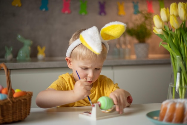 Um garotinho usando orelhas de coelho prepara ovos de Páscoa pintados para a Páscoa enquanto está sentado na mesa da cozinha Feliz Páscoa