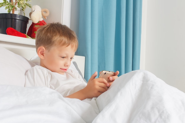 Um garoto de seis anos está descansando deitado na cama e segurando um smartphone nas mãos. Uma criança com um gadget em casa.