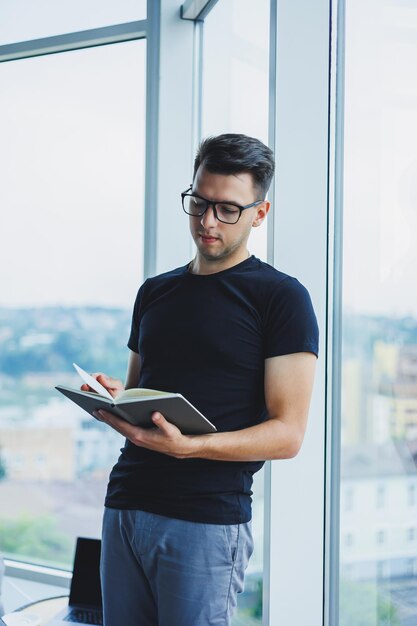 Um gerente estudantil fica perto de uma grande janela e lê notas em um caderno um jovem em uma camiseta preta com óculos fica no escritório perto da janela