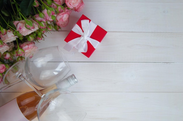 Um grande buquê de rosas cor de rosa um presente em uma caixa vermelha e uma garrafa de vinho rosé com copos deitar ao lado em um fundo branco de madeira com um lugar para texto