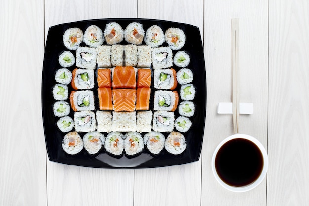 Um grande conjunto de sushi é colocado em um prato quadrado preto. Vista do topo. Mesa de madeira clara. Uma tigela de molho de soja e um pauzinho estão ao lado dela. Delicioso almoço ou jantar no estilo japonês. Foco suave.