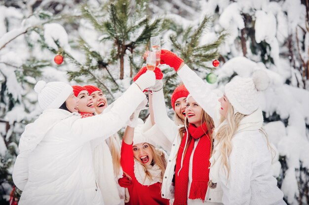 Um grande grupo de garotas com taças de champanhe nas mãos está na floresta de inverno. garotas com roupas vermelhas e brancas com bebidas de ano novo em uma floresta coberta de neve.