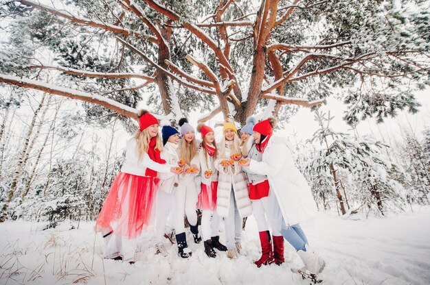 Um grande grupo de garotas com tangerinas está parado na floresta de inverno. garotas com roupas vermelhas e brancas com frutas em uma floresta coberta de neve.