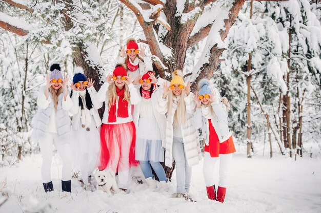 Um grande grupo de garotas com tangerinas está parado na floresta de inverno. Garotas com roupas vermelhas e brancas com frutas em uma floresta coberta de neve.