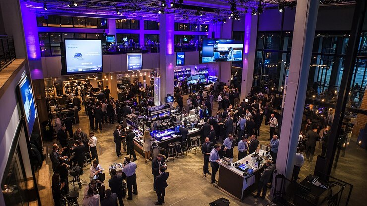 Foto um grande grupo de pessoas se reúne em um espaço de evento moderno com uma barra de várias telas exibindo visuais e um teto alto
