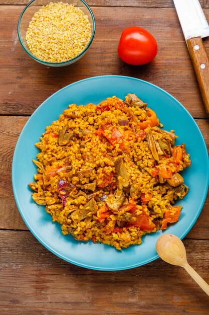 Foto um grande prato de bulgur com legumes e cogumelos em uma mesa ao lado de uma colher de pau