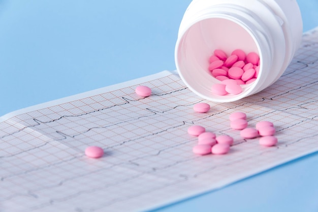 Um grande punhado de pílulas cor-de-rosa derramadas de uma jarra branca em um eletrocardiograma do coração em um fundo azul O conceito de estilo de vida saudável e exame médico oportuno