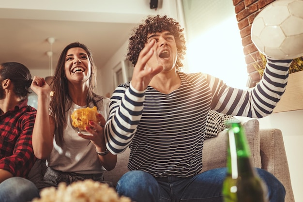 Foto um grupo de amigos assiste à transmissão de um evento esportivo. sentam-se em frente à tv da sala, comem lanche, bebem cerveja e torcem pelo time favorito.