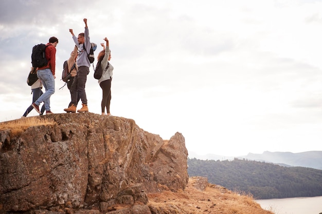 Foto um grupo de cinco amigos adultos felizes torce com os braços no ar no cume de uma montanha durante uma caminhada