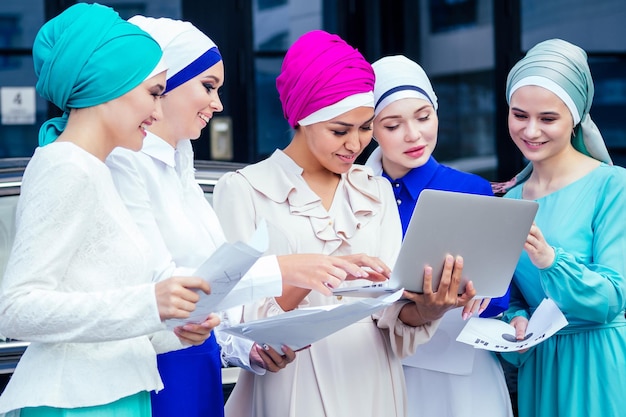 Um grupo de cinco mulheres de negócios jovens e atraentes em hijab multicolorido vestido muçulmano e um turbante cobrindo a cabeça trabalhando laptop e se comunicando e tendo trabalho de equipe e conceito de sucesso