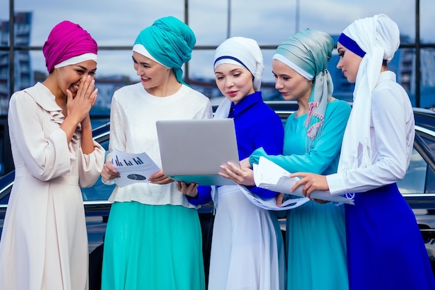 Um grupo de cinco mulheres de negócios jovens e atraentes em hijab multicolorido vestido muçulmano e um turbante cobrindo a cabeça trabalhando laptop e se comunicando e tendo trabalho de equipe e conceito de sucesso