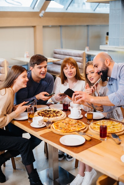 Foto um grupo de jovens amigos alegres está sentado em um café, conversando e tirando selfies ao telefone. almoço na pizzaria.