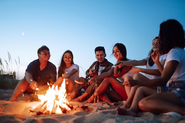 Um grupo de jovens amigos sentados na praia e fritando salsichas um homem está tocando guitarra tempo de acampamento