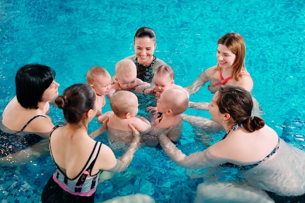Um grupo de mães com seus filhos pequenos em uma aula de natação infantil com um treinador.