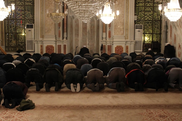 Um grupo de pessoas reza em uma mesquita, com candelabros pendurados no teto.