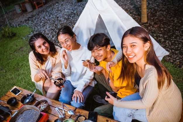 Um grupo de pessoas sorrindo juntas enquanto se sentam e comem carne grelhada no acampamento