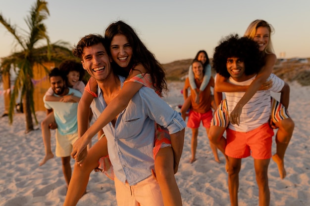 Um grupo multiétnico de amigos aproveitando o tempo juntos, caminhando na areia, homens carregando mulheres nas costas