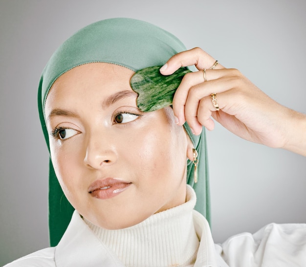 Um gua sha sendo usado no rosto de uma jovem muçulmana isolado contra o fundo cinza do estúdio Mulher vestindo um hijab ou lenço na cabeça usando uma ferramenta antienvelhecimento para reduzir rugas e promover a renovação celular