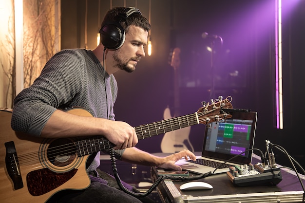 Um guitarrista com fones de ouvido conectados a um mixer de som enquanto grava o som, trabalha com um laptop em um programa de gravação.