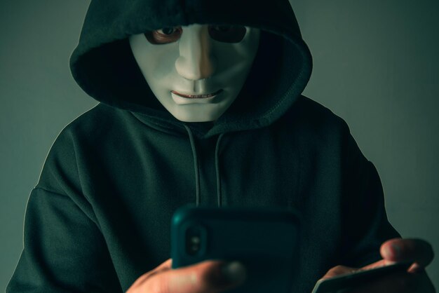 Um hacker mascarado anônimo está usando um smartphone para penetrar nas informações financeiras do cartão de crédito Conceito de hacking e malware