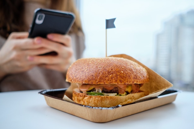 Um hambúrguer em uma mesa na frente de uma garota que está olhando para um smartphone.