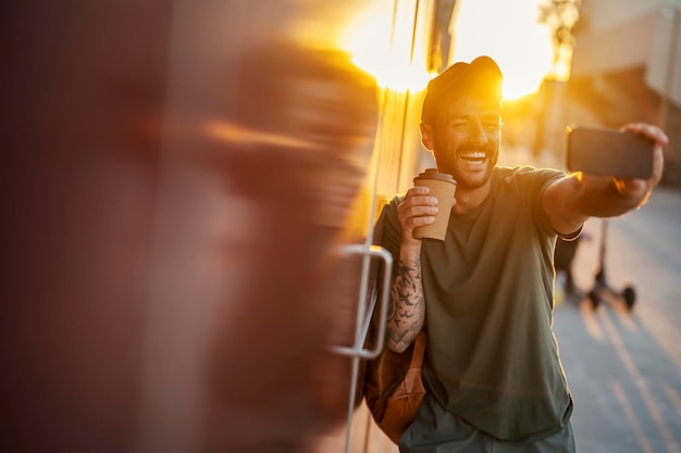 Um hipster feliz está tirando selfies na rua com seu café