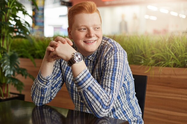 Um hipster ruiva positiva com sardas vestindo camisa xadrez elegante e assistir sentado à mesa de madeira no restaurante acolhedor