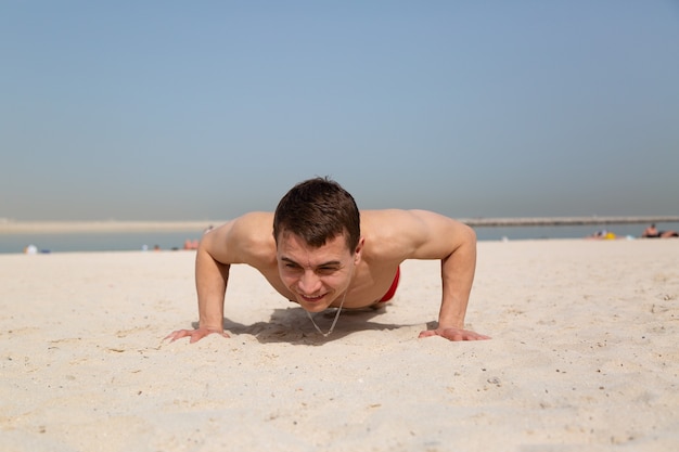 Um homem adulto, caucasiano, de 35 a 40 anos pratica esportes, flexões na areia sob o sol forte.