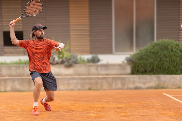 Um homem adulto de camiseta vermelha jogando tênis na quadra coloca a raquete no ar