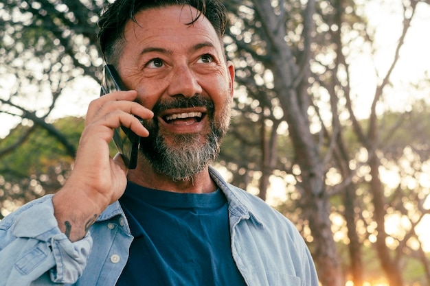 Um homem adulto feliz sorrindo e falando ao telefone ao ar livre no parque com a luz do pôr do sol e árvores no fundo Homens casuais de jeans usando celular para ligar para amigos Retrato alegre de pessoa
