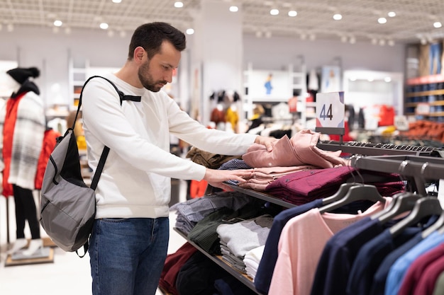 Um homem adulto moreno bonito em um moletom branco escolhe uma camisa da moda elegante em uma loja de roupas em